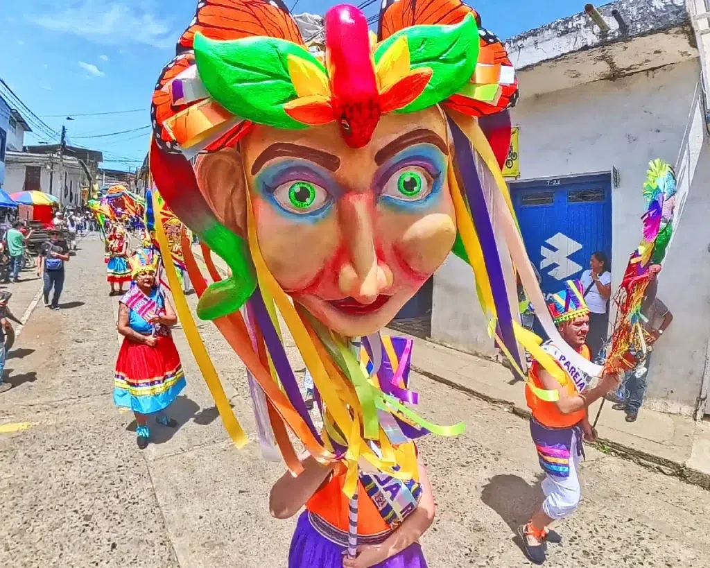 Mascarón gigante y participantes con trajes folclóricos coloridos desfilando por las calles de Quinchía durante las Fiestas de La Villa de Los Cerros.